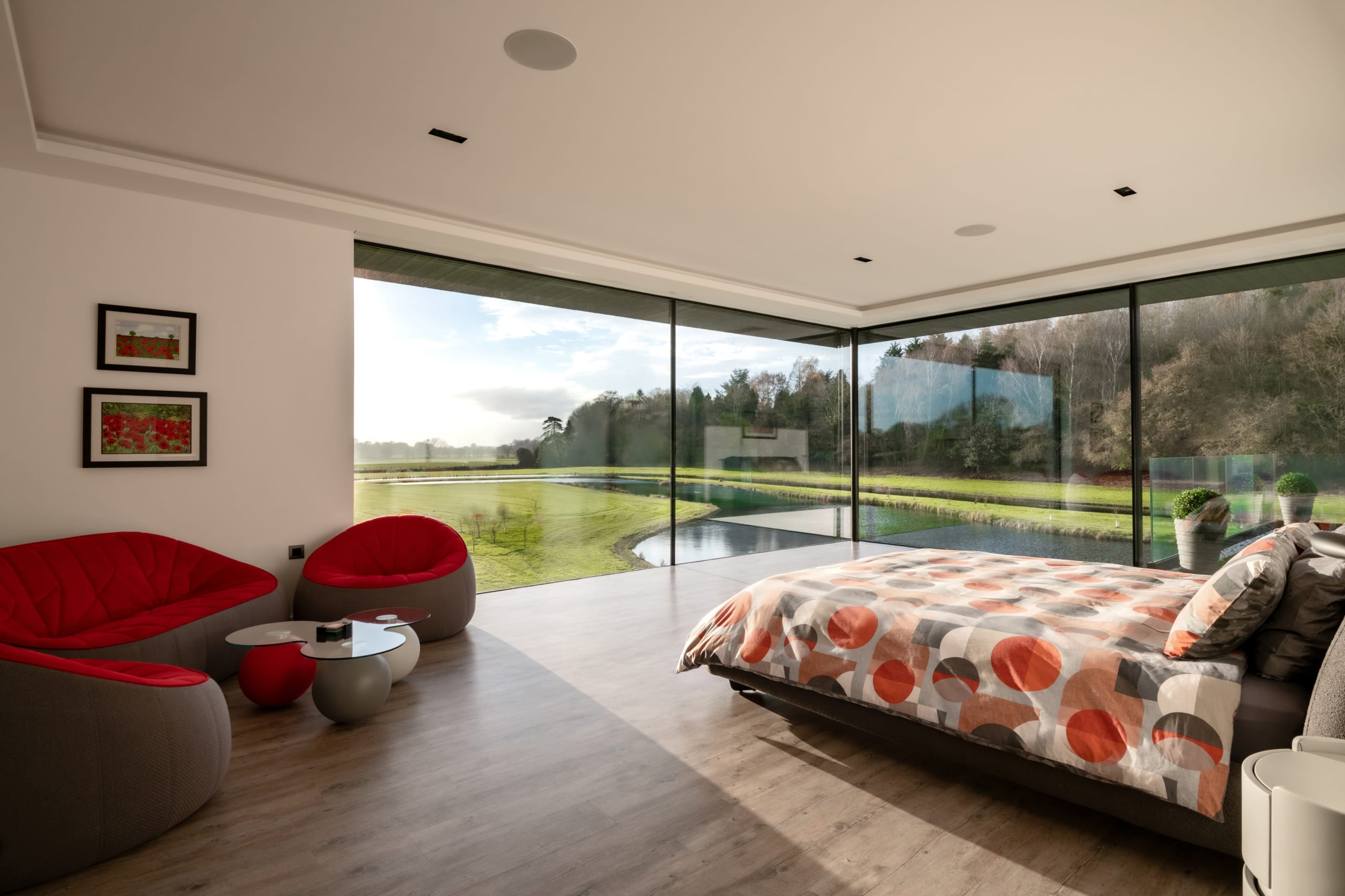 Modern bedroom with floor-to-ceiling glass walls overlooking landscaped grounds, featuring red accent chairs and geometric bedding